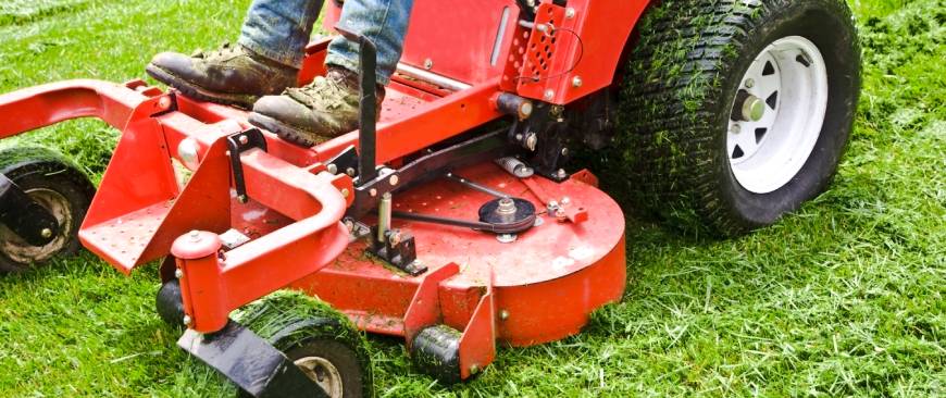 Close-up of a man’s legs riding a big red lawn mower with big pneumatic tires. The mower is cutting green grass outdoors.