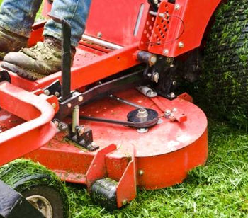 Close-up of a man’s legs riding a big red lawn mower with big pneumatic tires. The mower is cutting green grass outdoors.