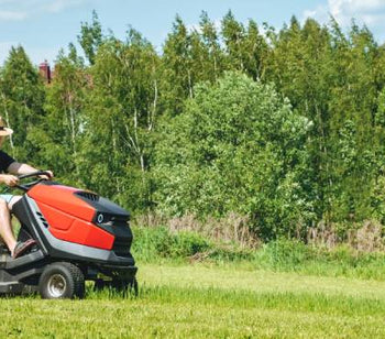 A man wearing a hat, a T-shirt, shorts, and ear protection riding a red lawn mower in a field with trees behind him.