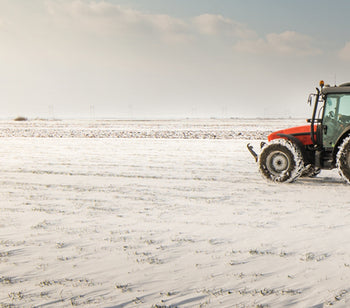 A red tractor moves over flat land in the winter. There is snow on the ground and it is a sunny day with clouds in the sky.