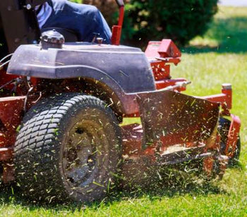 A person is riding a red lawn mower with large tires and mowing grass on the side of a road. The sun is shining outside.