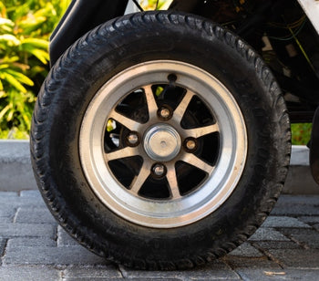 A golf cart wheel is attached to a white golf cart. There are plants and flowers nearby, and it's a sunny day.