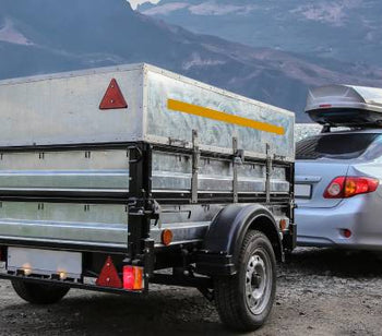 A light gray sedan with an attached trailer is parked on a rocky surface overlooking a body of water and mountains.