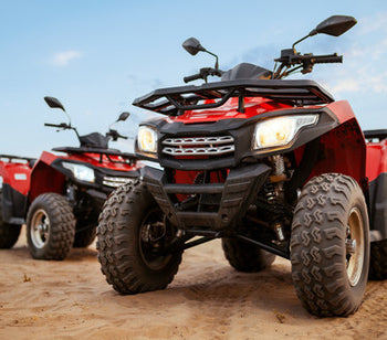 Two red ATVs parked on a sandy surface. There are tire tread marks in the sand, and the sky has a few clouds.