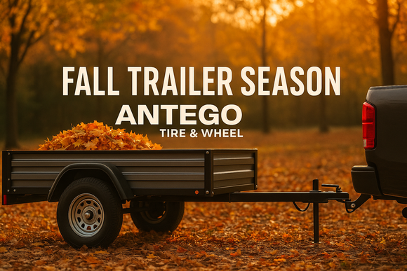 Fall landscape with utility trailer loaded beside a truck, showing two trailer wheels with ST tires, autumn leaves on the ground, and a warm orange sky—blog hero for Antego Tire & Wheel about fall trailer season.