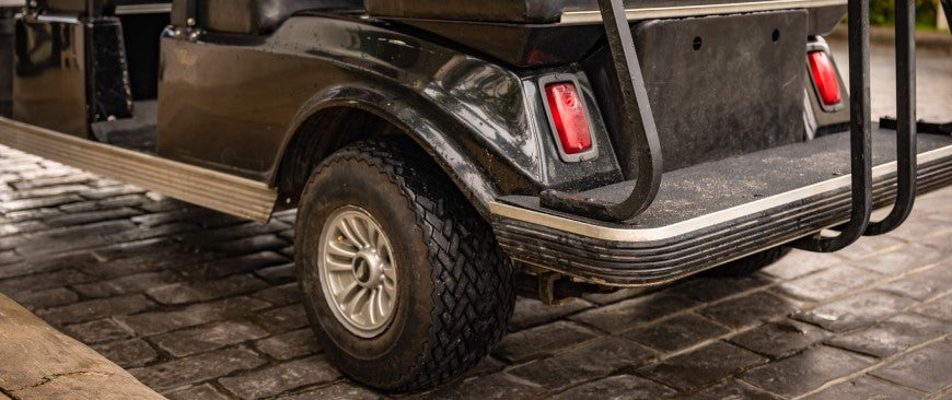 Close up of the left backside of a black painted golf cart with thick rubber tires, a metal wheel, and small stop lights parked outdoors